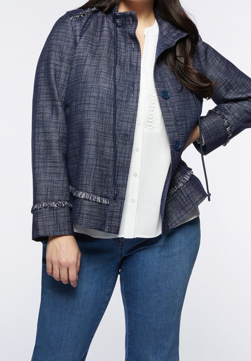 Marinblå texturerad jacka med fransad kant, knappar fram och dragsko. Bärs över en vit topp, kombinerad med blå jeans.