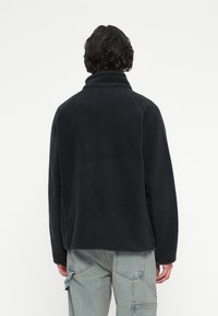 Persona con capelli scuri e mossi di spalle, che indossa una giacca in pile nera e pantaloni cargo in denim azzurro chiaro su uno sfondo neutro.
