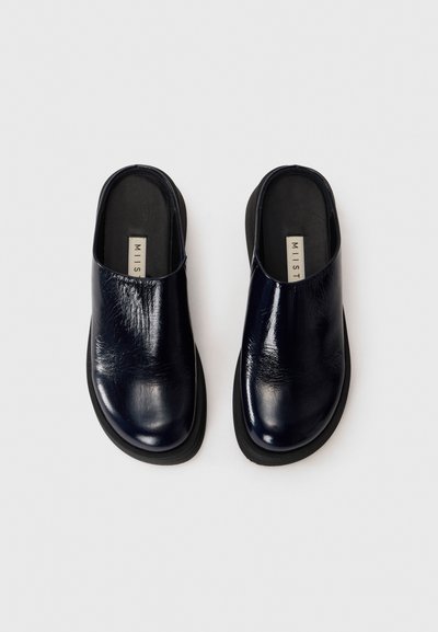 Mornarske modre usnjene čevlje s sijajno zaključkom, zaobljenim prstom in nizkim črnim gumijastim podplatom; imajo odprto zadaj in minimalističen dizajn.