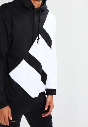 Mann trägt schwarzen und weißen Hoodie mit diagonaler geometrischer Gestaltung und Kordelzug, kombiniert mit zerrissenen schwarzen Jeans, steht vor weißem Hintergrund.