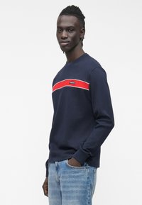 Tumesinise varrukaga triiksärk soonilise tekstuuriga, millel on horisontaalne punase ja valge triip rinnal ning väike brändilt pärit märk.