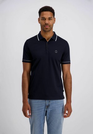 Mann steht mit Blick nach vorne, trägt ein schwarzes Poloshirt mit weißem Saum und hellblaue Jeans vor einem schlichten Hintergrund.