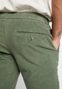 Close-up van een persoon in groene corduroy broek met een knoopzak op de achterkant en de rechterhand in de zijzak, zichtbaar is een beige geribbelde top.
