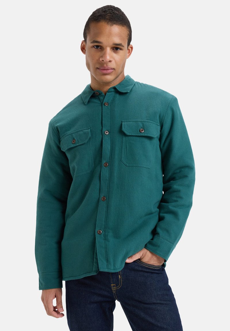 Szmaragdowa polarowa koszula z guzikami i kołnierzykiem, z dwoma kieszeniami na piersi oraz brązowymi guzikami, w połączeniu z ciemnoniebieskimi dżinsami. Gładka faktura i swobodny styl.