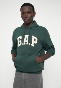 Zielona bluza z kapturem z kremowym logo "G.A.P." na piersi, kieszenią typu kangur oraz ściągaczami na rękawach. Materiał wydaje się miękki i wygodny.