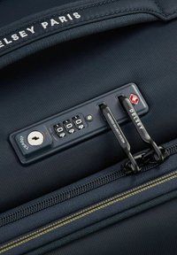 Detalle de maleta azul marino que incluye un candado de combinación, un agujero para llave y cremalleras negras con costuras amarillas en contraste; marcada con "Delsey Paris."