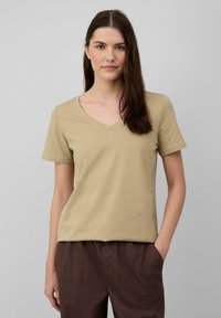 Beige V-Ausschnitt-T-Shirt aus leichtem Baumwollmaterial, mit kurzen Ärmeln und lockerer Passform. Kombiniert mit braunen Hosen, vor einem neutralen Hintergrund.