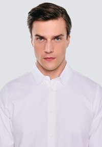 Biała koszula zapinana na guziki z teksturowanej tkaniny, klasycznym kołnierzykiem i widocznymi guzikami z przodu, noszona przez modela na neutralnym tle.