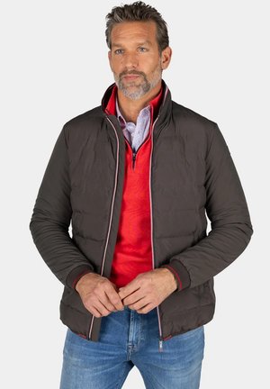 Man met baard die een zwart gewatteerd jack draagt over een rode trui met een kwart-ritssluiting en blauwe jeans, staand met gevouwen handen.