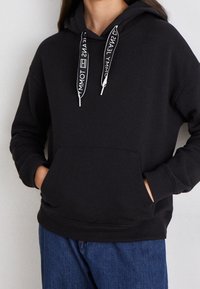 Black hooded sweatshirt with white branded drawstrings, front pocket, and ribbed cuffs. Casual fit with soft, textured fabric.