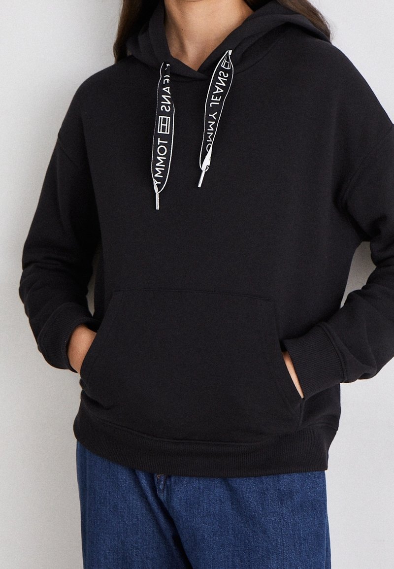 Black hooded sweatshirt with white branded drawstrings, front pocket, and ribbed cuffs. Casual fit with soft, textured fabric.