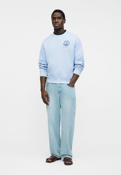 Άνδρας που στέκεται με το ένα χέρι στην τσέπη, φοράει ανοιχτό μπλε φούτερ, χαλαρά τζιν ανοιχτού χρώματος και μαύρα σανδάλια σε απλό φόντο.