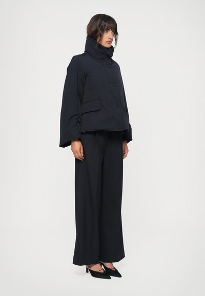 Zwarte cropped pufferjack met hoge kraag, grote knopen en een voorkantzak, gecombineerd met wijde zwarte broek en zwarte hakken.
