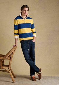 Jeune homme portant un maillot de rugby rayé jaune et bleu, un jean foncé et des baskets marron, appuyé sur une chaise en osier devant un fond beige.