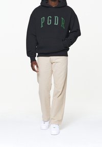 Felpa nera con logo "PGDR" in verde, dotata di una tasca frontale. Abbinata a pantaloni beige chiaro e sneakers bianche. Vestibilità standard.