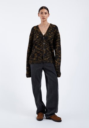 Schwarz-gelb gesprenkelte Strickjacke mit V-Ausschnitt, sechs Knöpfen und langen Ärmeln, kombiniert mit dunkelgrauen, weit geschnittenen Jeans und braunen Schuhen.