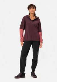 Giacca impermeabile bordeaux e rosa con cappuccio, chiusura a zip e tasche laterali; abbinata a pantaloni neri e scarpe scure.