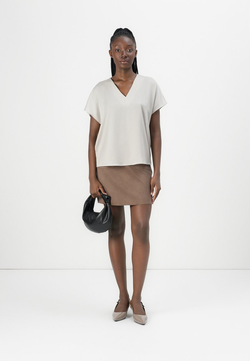 Top de manga corta en beige claro con escote en V, combinado con una falda corta marrón, sosteniendo un bolso negro y luciendo unos zapatos de punta bicolor.