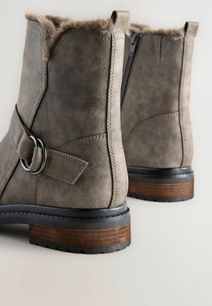 Grå anklar stövlar med en mjuk pälsfoder, lädermaterial, rund tå, dragkedjestängning och en staplad träklack; har ett dekorativt rem.