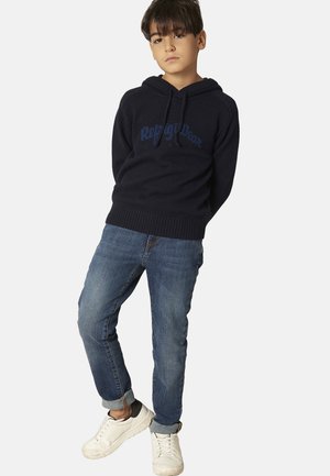 Felpa lavorata a maglia blu navy con logo prominente, polsini e orlo a costine. Abbinata a jeans azzurri e sneakers bianche con accenti neri.