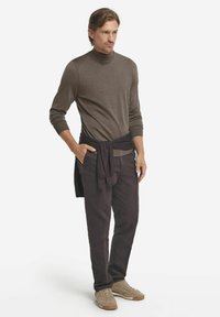 Dunkelbrauner Rollkragenpullover, kombiniert mit dunklen Hosen und beigen Sneakers. Pullover lässig um die Taille gebunden, glatte Textur.