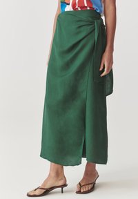 Falda envolvente verde hecha de un tejido ligero, con una abertura lateral, detalle de cintura atada y textura suave. Se lleva con sandalias marrones.