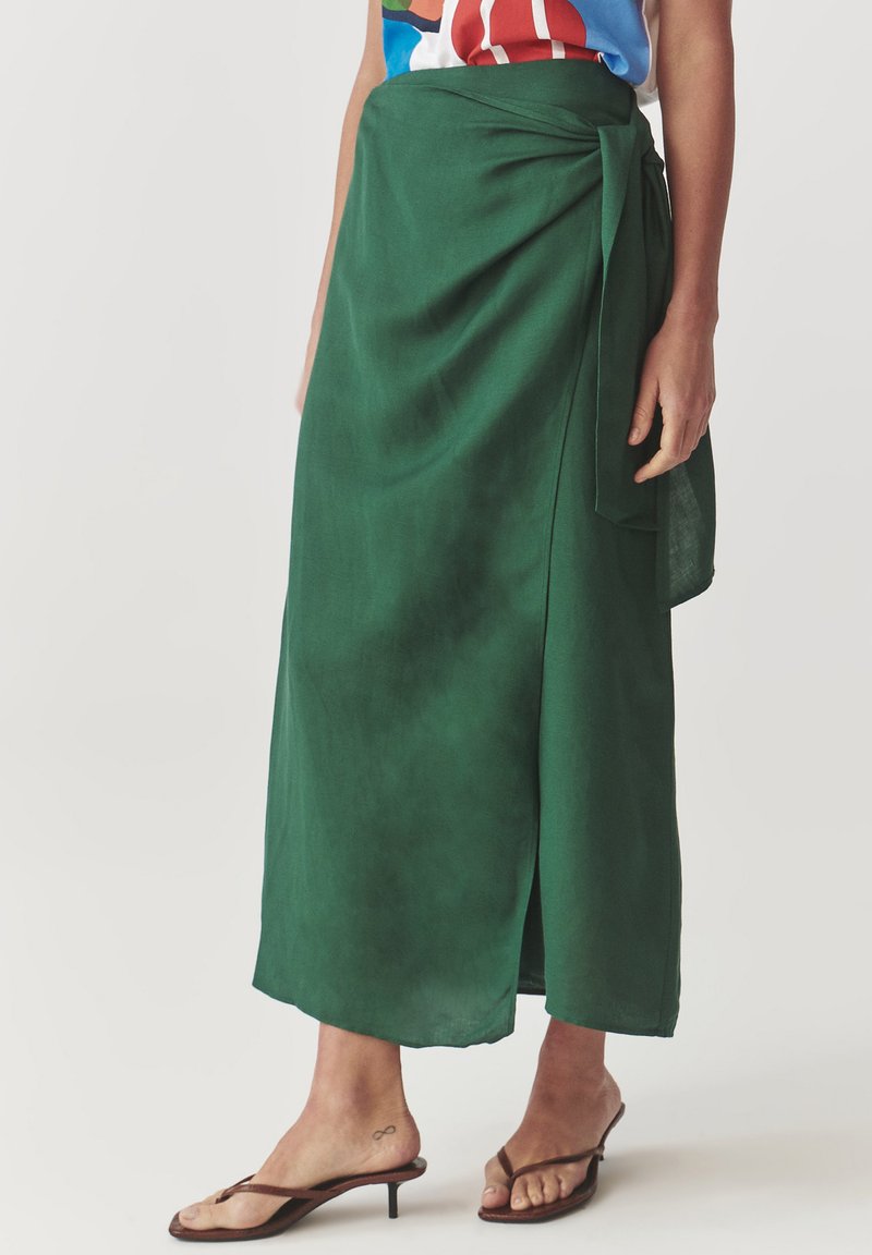 Falda envolvente verde hecha de un tejido ligero, con una abertura lateral, detalle de cintura atada y textura suave. Se lleva con sandalias marrones.