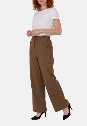 Donna con capelli ricci rossi che indossa una t-shirt bianca, pantaloni marroni a vita alta a gamba larga con bottoni laterali e scarpe nere con tacchi alti, in piedi.