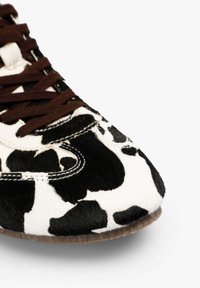 Zapatilla con estampado de vaca en blanco y negro, con cordones marrones, parte superior de textura suave y suela de goma. Detalle de costura expuesta en el lateral.