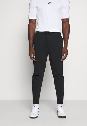 Uomo che indossa una maglietta bianca Nike, pantaloni jogger neri con coulisse, calzini bianchi Nike e sneakers bianche Nike, in piedi contro uno sfondo semplice.