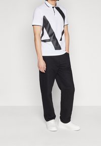 Biała koszulka polo z czarnym gumowanym wzorem graficznym, czarnymi detalami kołnierza i rękawów, zestawiona z luźnymi czarnymi spodniami i białymi trampkami.