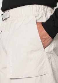 Pantaloni corti beige chiaro con vita elastica e tasca laterale. Presentano un sottile logo "Calvin Klein Jeans" in carattere bianco sul lato sinistro.