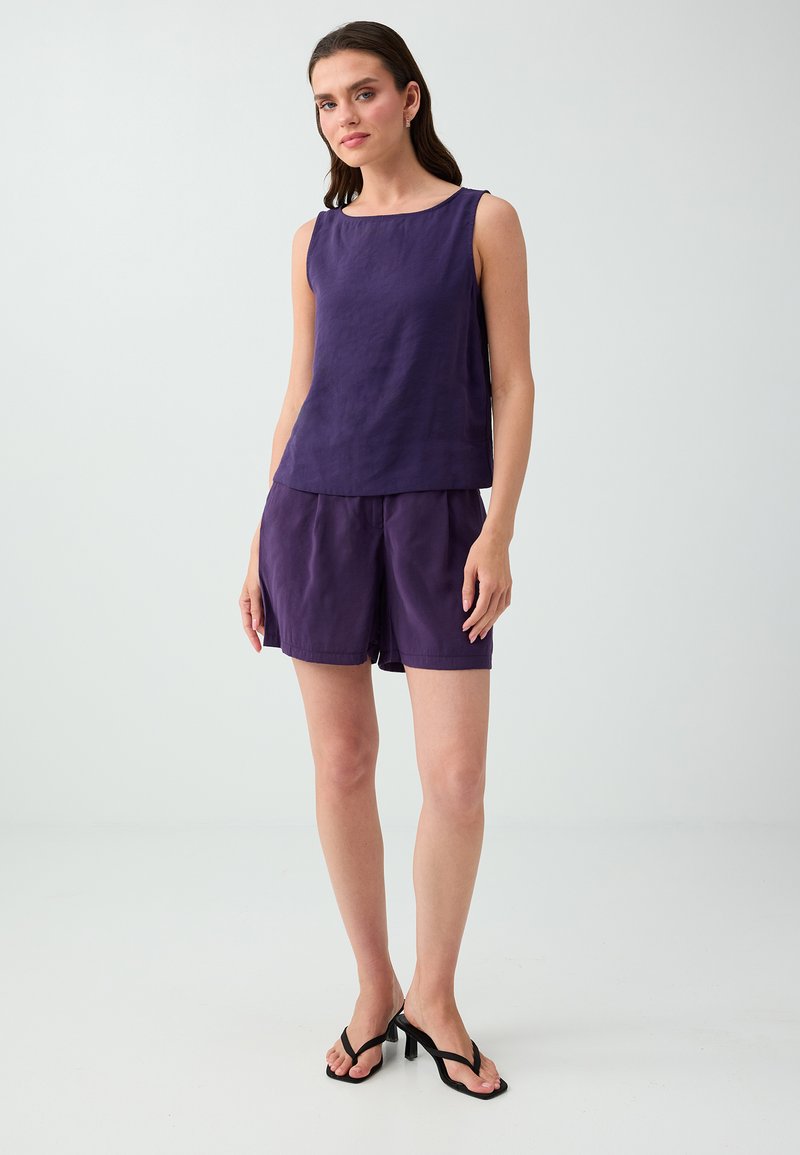 Femme debout portant un haut violet sans manches, un short assorti et des sandales à talons noires sur un fond clair uni.