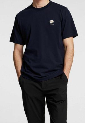 T-shirt en coton bleu marine à manches courtes, avec un petit motif beige clair sur la partie supérieure gauche de la poitrine. Design simple.