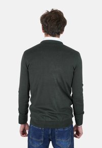 Maglione verde scuro a maglia con scollo rotondo e polsini a costine, abbinato a jeans blu. Il tessuto appare liscio e leggermente strutturato.