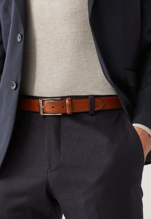 Uomo con blazer blu navy, maglione beige, pantaloni scuri e cintura di pelle marrone con fibbia in ottone, mano in tasca.