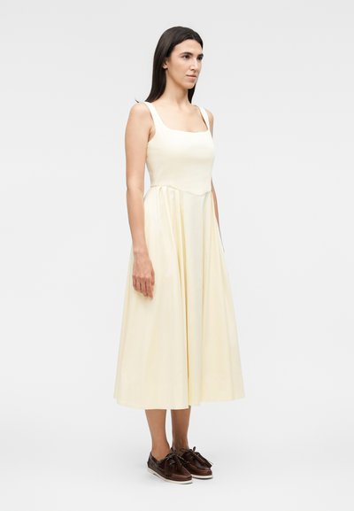 Γυναίκα με μακριά σκούρα μαλλιά που φοράει αμάνικο κρεμ midi φόρεμα και καφέ δερμάτινα παπούτσια, στέκεται μπροστά σε ένα απλό φωτεινό φόντο.