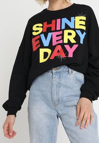 Black sweatshirt with oversized fit, featuring colorful text "SHINE EVERY DAY" in bold letters. Lightweight, ribbed cuffs, and relaxed design.