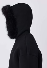 Veste noire à capuche avec une épaisse bordure en fourrure noire le long du bord de la capuche, vue de côté sur un fond uni.