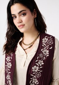 Bordeauxkleurig doorgestikt vest met witte bloemenborduursels, gedragen over een crèmekleurige, getextureerde top, gecombineerd met een meerstrengige ketting.