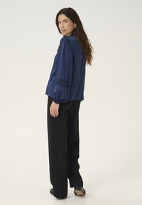 Blouse bleu marine à manches longues avec épaulettes froncées et dentelle, associée à un pantalon noir à jambes larges. Texture de tissu lisse.