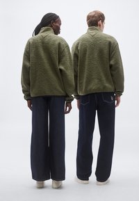 Fleecepullover i olivengrøn, tekstureret overflade, høj krave, afslappet pasform. Bæres med mørkeblå viddebukser og hvide sneakers.