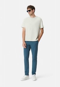 Jasnoszary t-shirt z krótkim rękawem, dopasowane spodnie w kolorze morskim oraz białe trampki. Osoba w ciemnych okularach przeciwsłonecznych, stojąca z rękami w kieszeniach.