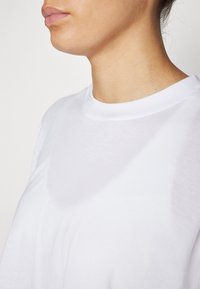 T-shirt bianco in cotone con scollo rotondo e maniche corte. Il tessuto appare liscio con una leggera lucentezza ed è leggermente traslucido.