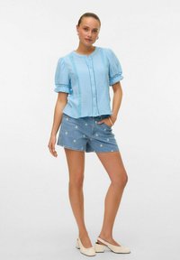 Blusa azzurra chiara con texture, maniche a sbuffo e dettagli in pizzo, abbinata a pantaloni in denim con ricami floreali e scarpe slip-on bianche.