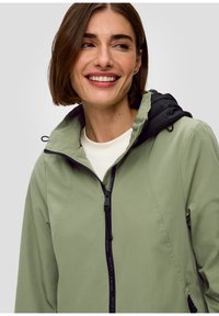 Veste légère verte avec une capuche noire, dotée d'une fermeture éclair intégrale, d'un design épuré et d'une texture lisse. Idéale pour un usage casual.