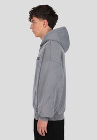 Sivi pulover s kapuco z velikim dizajnom, teksturirano tkanino, rebrastimi manšetami in kangaroo žepom, pogled od strani.