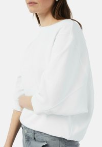 Witte sweatshirt met een ontspannen pasvorm, wijde mouwen en geribde manchetten. Gepaard met grijze high-waisted broeken, met een knoopdetail.