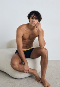 Calzoncillos negros con detalle amarillo en la cinturilla, modelo sentado en una silla texturizada de color beige, con un fondo neutro y una paleta de colores claros.