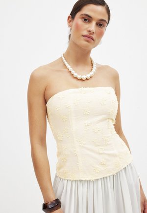 Beżowa bluzka bez ramiączek z kwiatowym haftem, dopasowany fason, teksturowany materiał, zestawiona z naszyjnikiem z pereł i szarą plisowaną spódnicą.